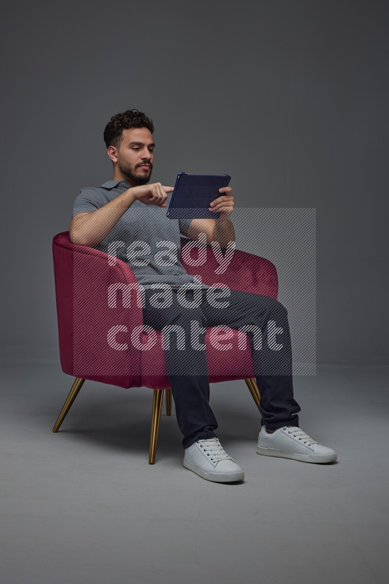 A man wearing casual and using his tablet while sitting on a burgundy chair eye level on a gray background