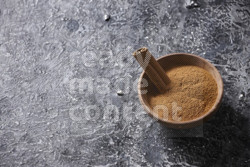 Wooden bowl full of cinnamon powder and a cinnamon stick on a textured black background