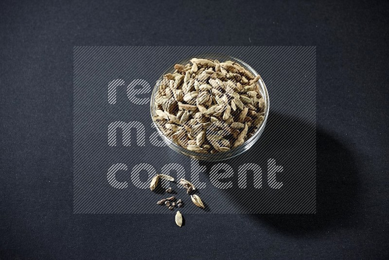 A glass bowl full of cardamom and more seeds spreaded beneath the bowl on black flooring