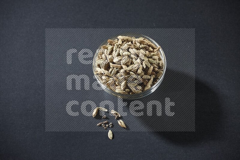 A glass bowl full of cardamom and more seeds spreaded beneath the bowl on black flooring