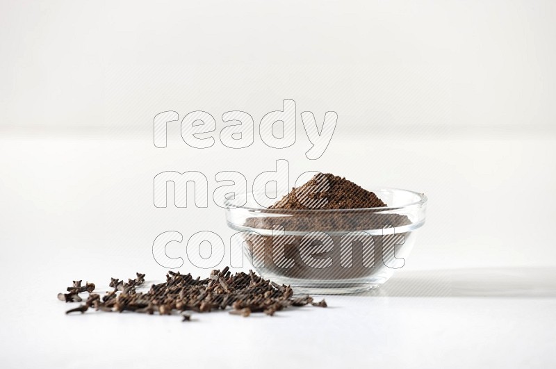 A glass bowl full of cloves powder and cloves grains spread on white flooring