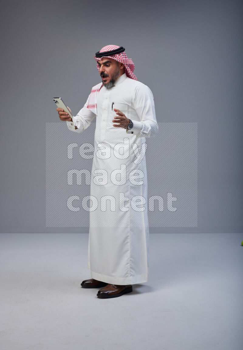 Saudi man Wearing Thob and red Shomag standing texting on phone on Gray background