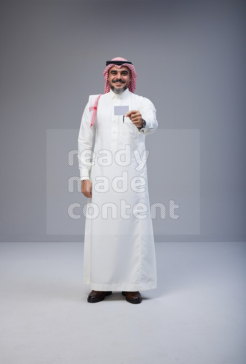 Saudi man Wearing Thob and red Shomag standing holding ATM card on Gray background