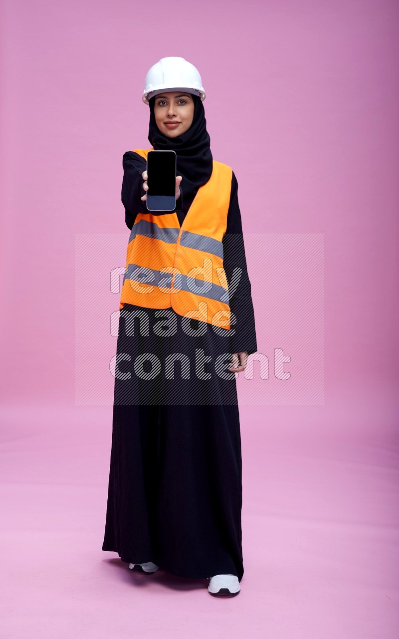 Saudi woman wearing Abaya with engineer vest and helmet standing showing phone to camera on pink background