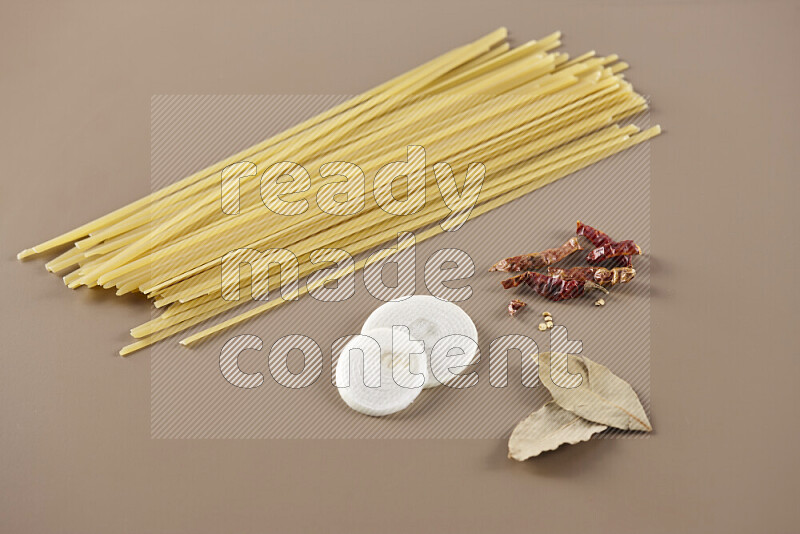 Raw pasta with different ingredients such as cherry tomatoes, garlic, onions, red chilis, black pepper, white pepper, bay laurel leaves, rosemary and cardamom on beige background