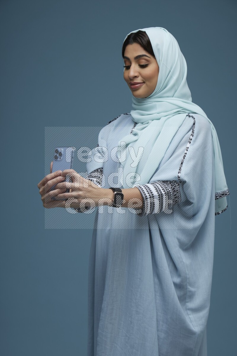 A Saudi woman Taking a Selfie on a blue background wearing a blue Abaya with hijab