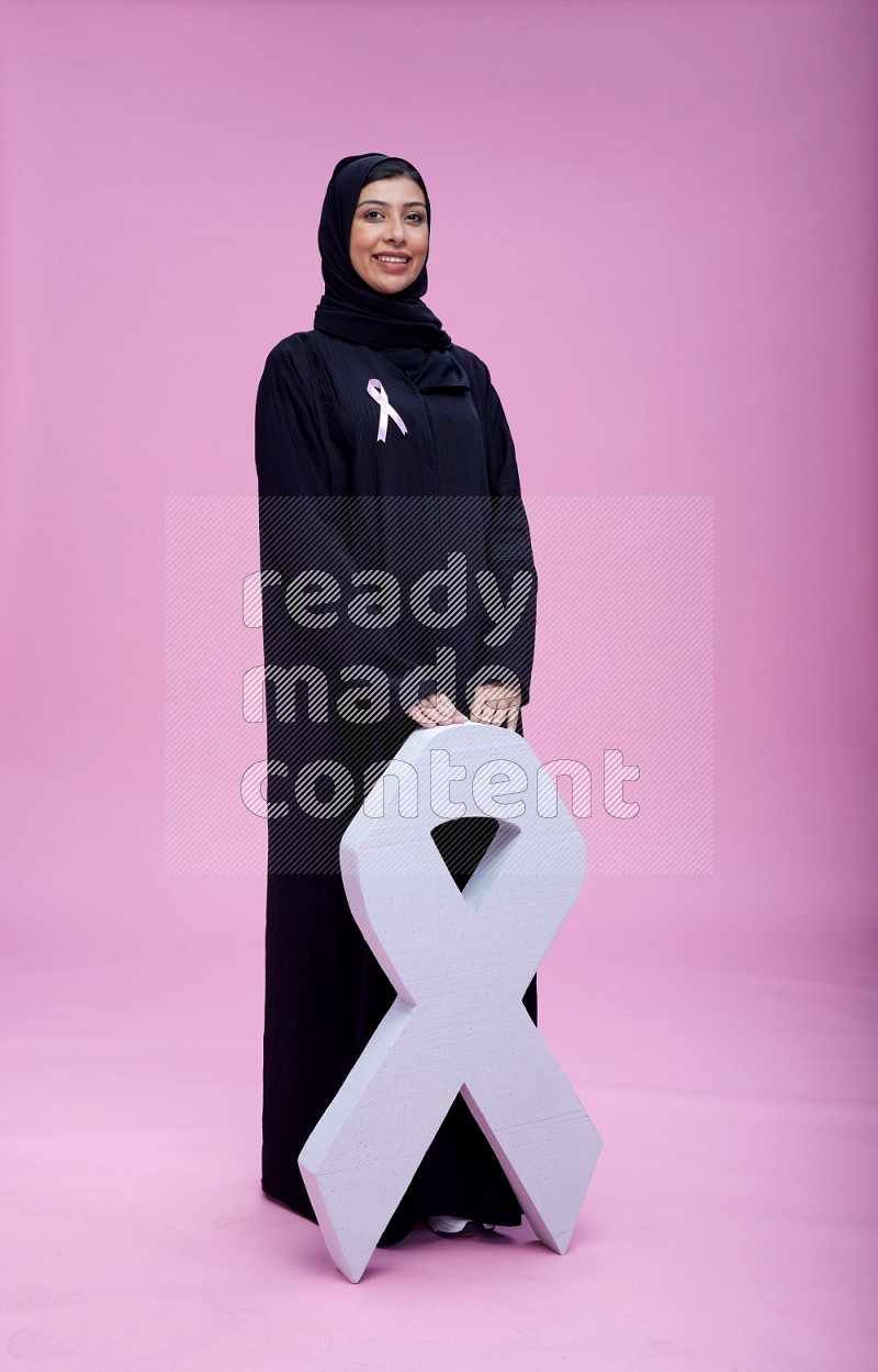 Saudi woman wearing pink ribbon on abaya standing holding awareness ribbon on plain pink background