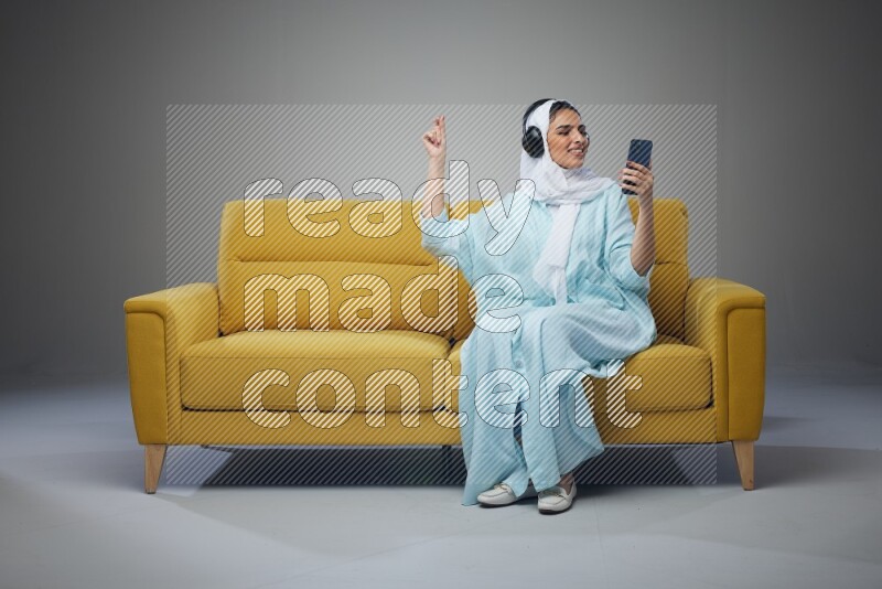 A Saudi woman wearing a light blue Abaya and a white head scarf sitting on a yellow sofa and holding an iPad while wearing headphone eye level on a grey background