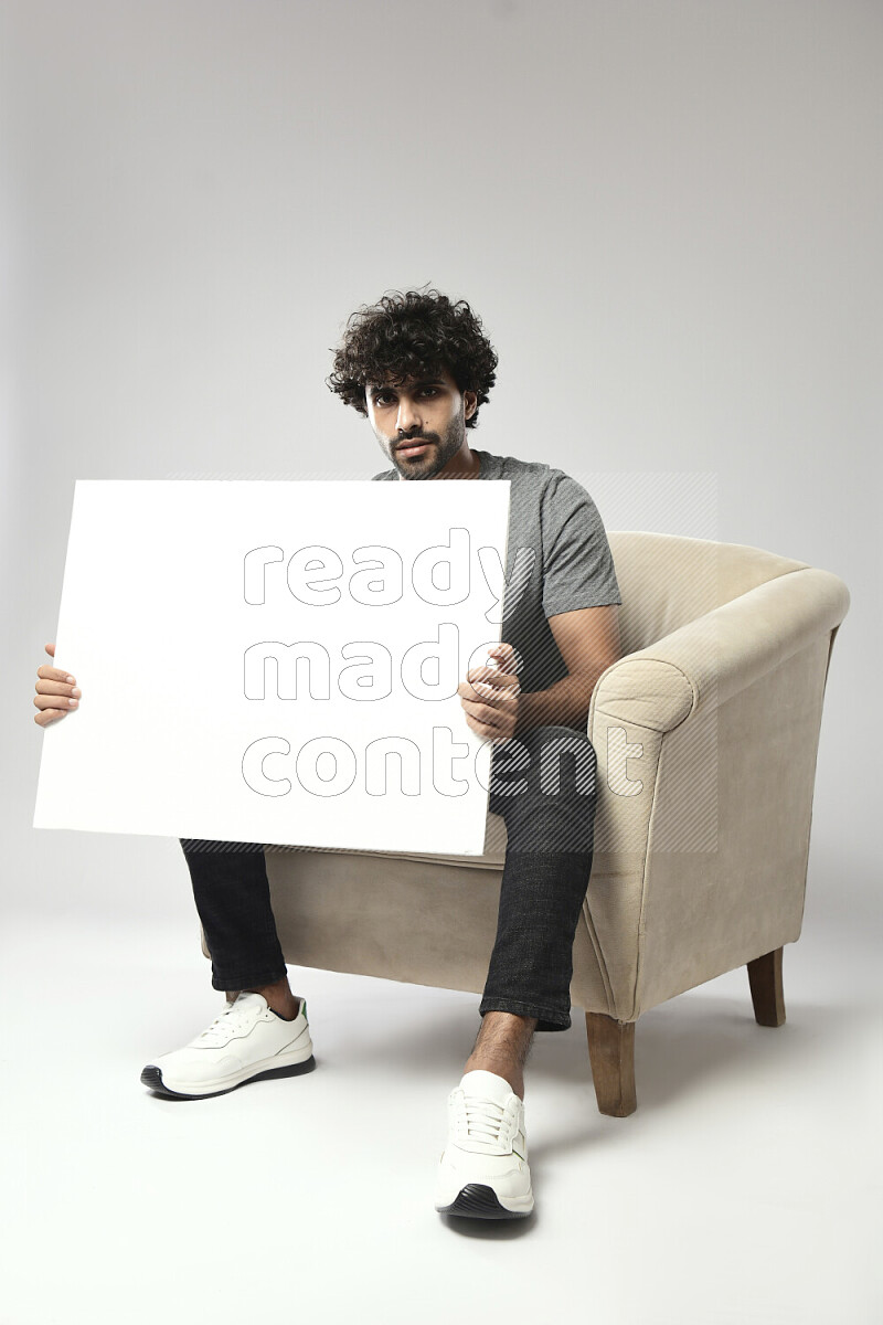 رجل يرتدي ملابس كاجوال يجلس علي كرسي و يحمل لوحة بيضاء علي خلفية بيضاء