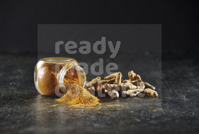 وعاء زجاجي للتوابل ممتلئ ببودرة الكركم مقلوب والبودرة تناثرت منه مع أصابع الكركم المجففة على خلفية سوداء