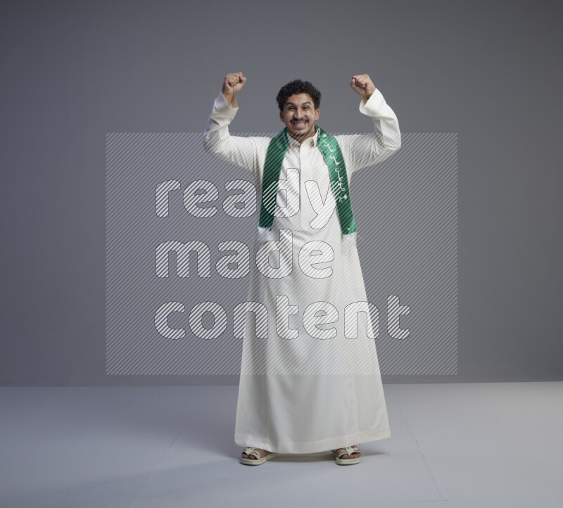A Saudi man standing wearing thob and Saudi flag scarf on gray background