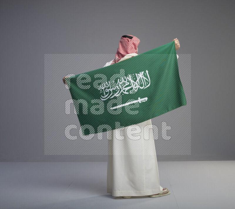 A saudi man standing wearing thob and red shomag holding big saudi flag on gray background