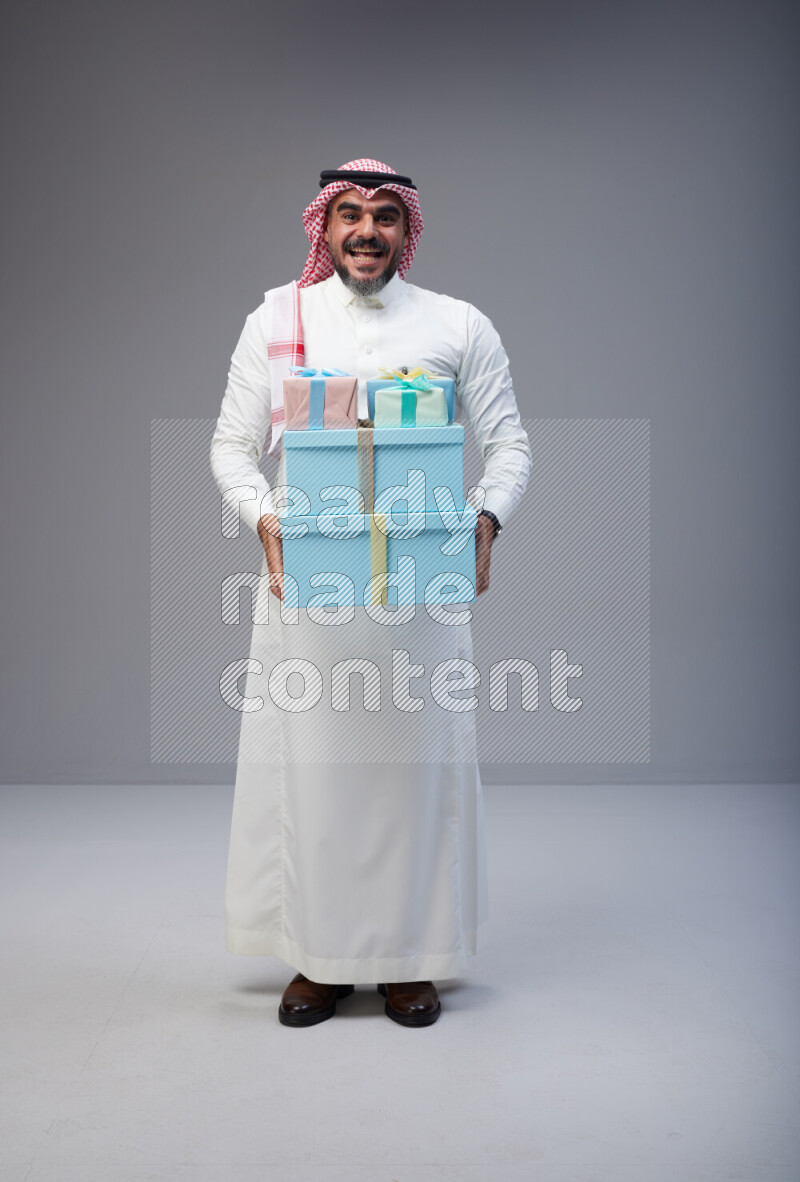 Saudi man Wearing Thob and red Shomag standing holding gift box on Gray background