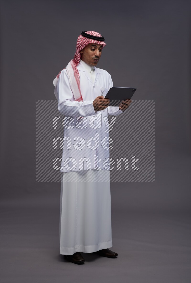 Saudi man wearing thob with lab coat and shomag with pocket employee badge standing working on tablet on gray background