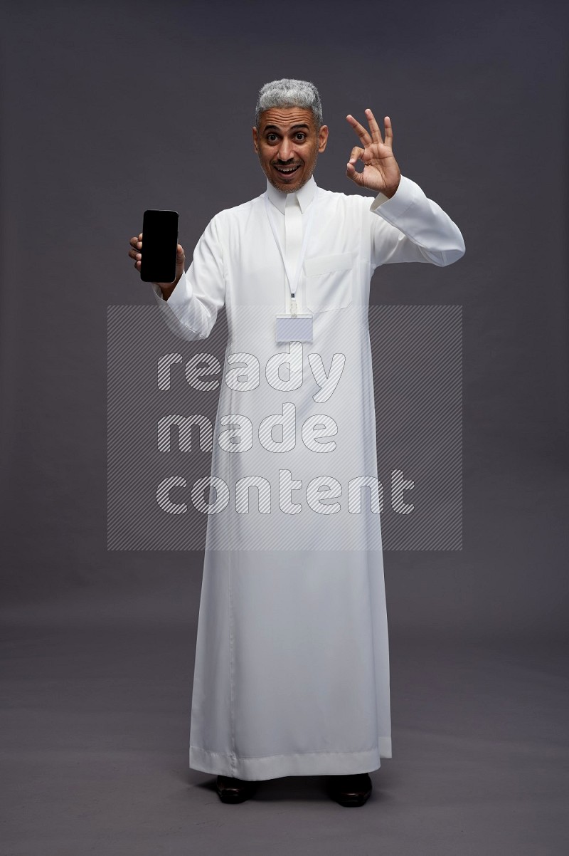 Saudi man wearing thob with neck strap employee badge standing showing phone to camera on gray background