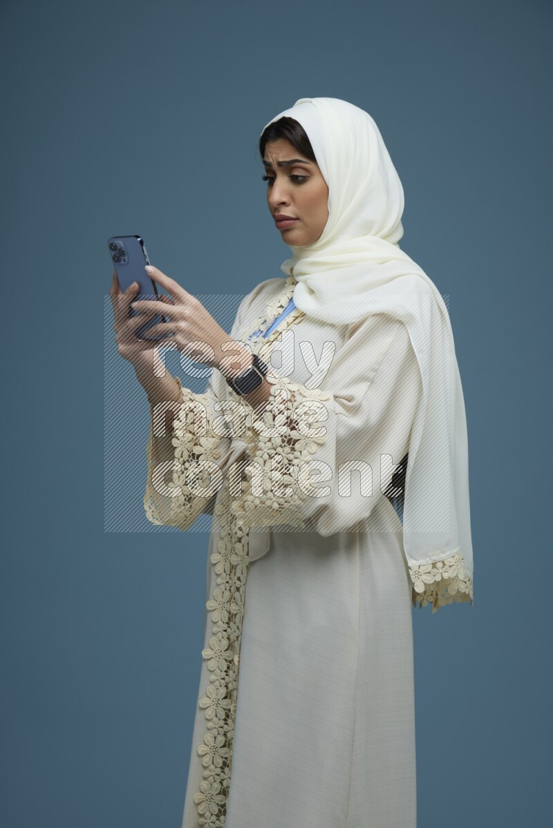 A Saudi woman Texting in a blue background wearing an off-white Abaya Hijab