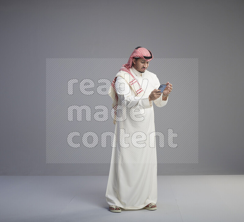 A  Saudi man standing wearing thob and red shomag texting on phone on gray background