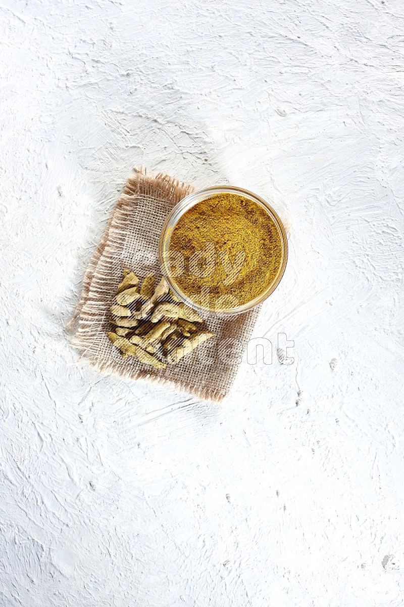 A glass bowl full of turmeric powder and dried turmeric whole finger on a piece of burlap on a textured white flooring