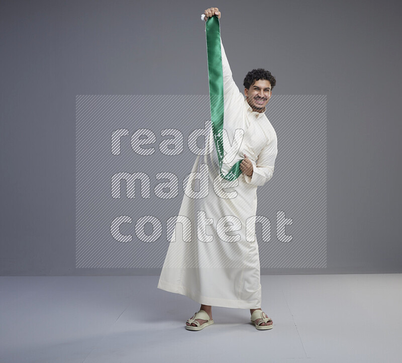 A Saudi man standing wearing thob holding Saudi flag scarf on gray background