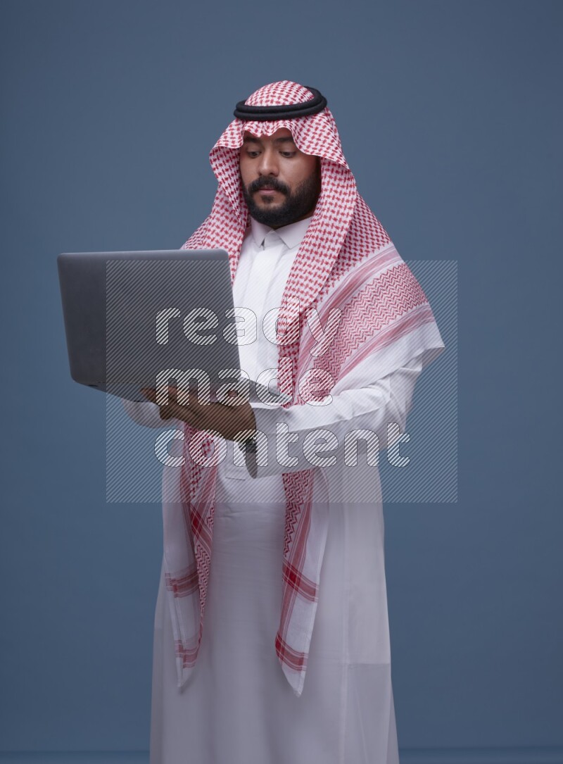 A man standing  with a laptop on Blue Background wearing Saudi Thob and Shomag