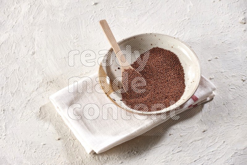 A multicolored pottery plate full of garden cress seeds with a wooden spoon full of the seeds on a napkin on a textured white flooring
