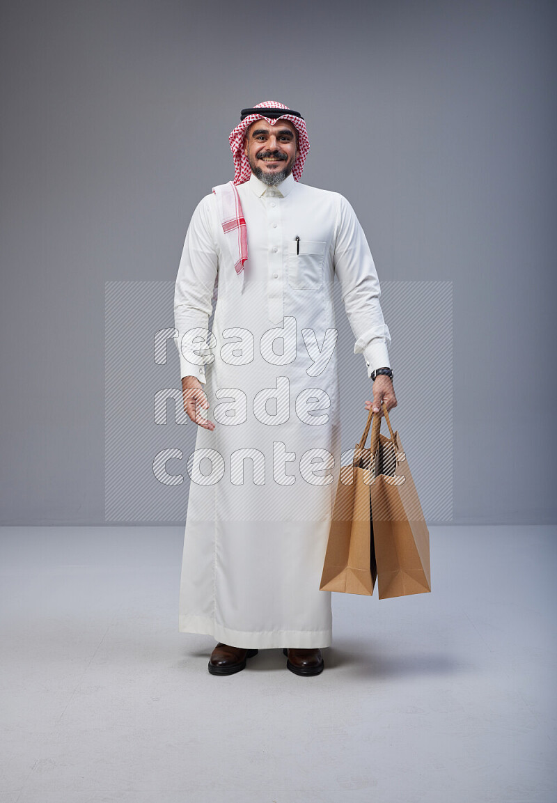 Saudi man Wearing Thob and red Shomag standing holding shopping bag on Gray background