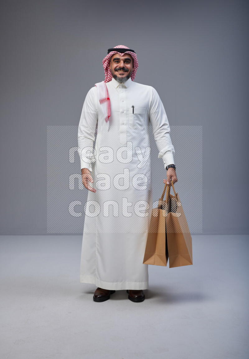 Saudi man Wearing Thob and red Shomag standing holding shopping bag on Gray background