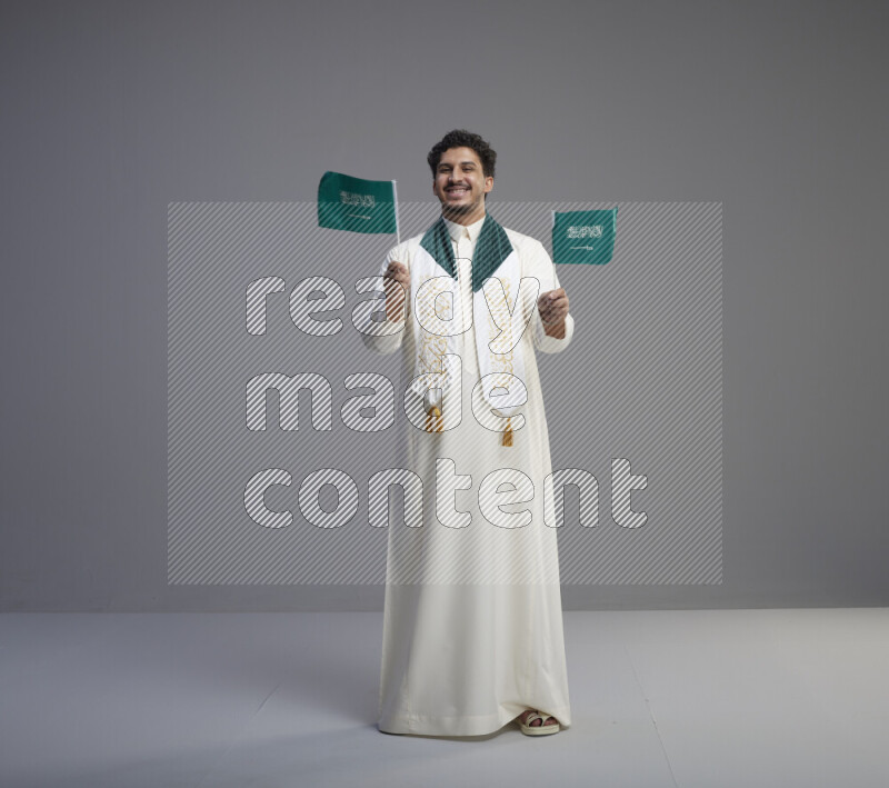 A Saudi man standing wearing thob and saudi flag scarf and holding small saudi flag on gray background