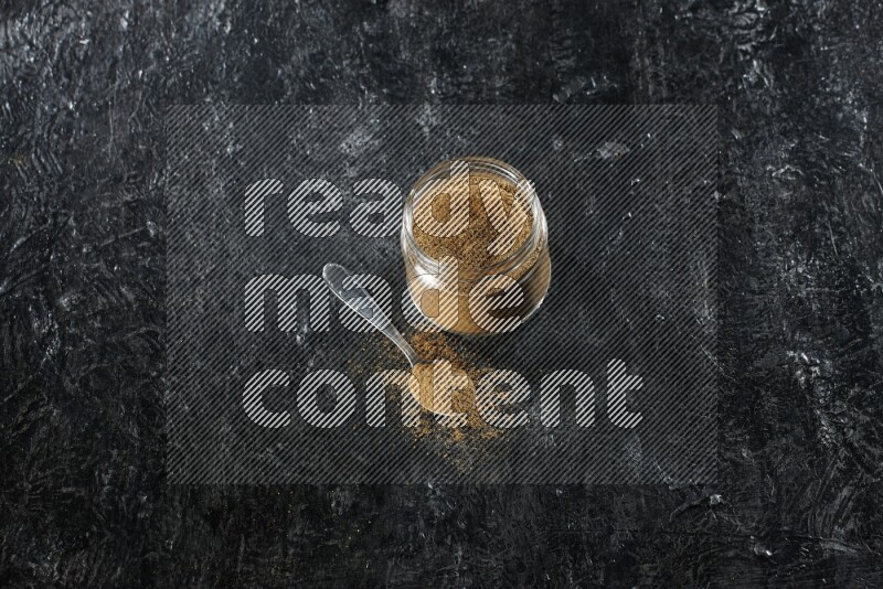 وعاء زجاجي للتوابل وملعقة معدنية ممتلئين ببودرة الكمون على خلفية سوداء