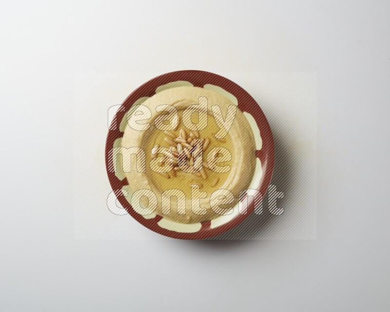 Hummus in a traditional plate garnished with pine nuts on a white background