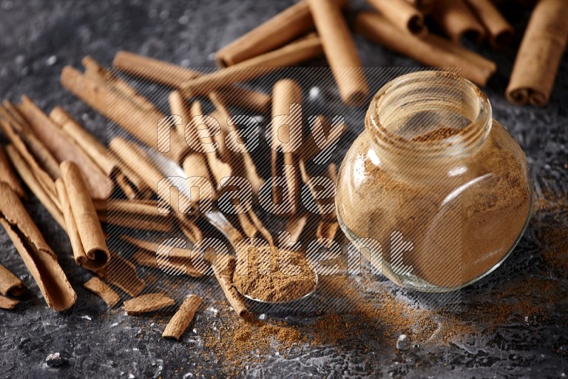 Herbal glass jar and a metal spoon full of cinnamon powder surrounded by cinnamon sticks on textured black background