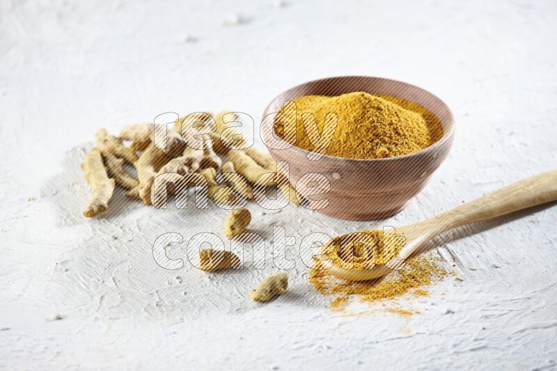 A wooden bowl and wooden spoon full of turmeric powder with dried turmeric fingers beside it on textured white flooring