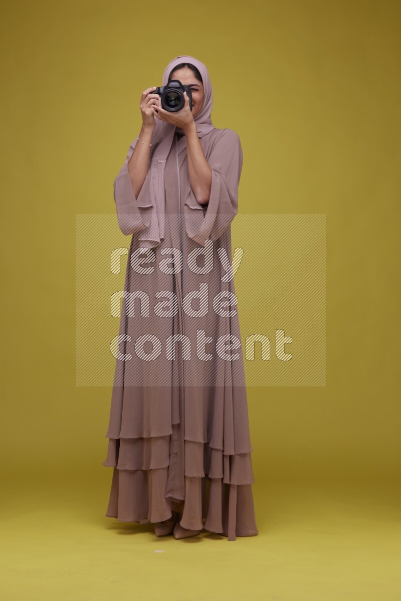 A woman Holding a Camera on a Yellow Background wearing Brown Abaya with Hijab