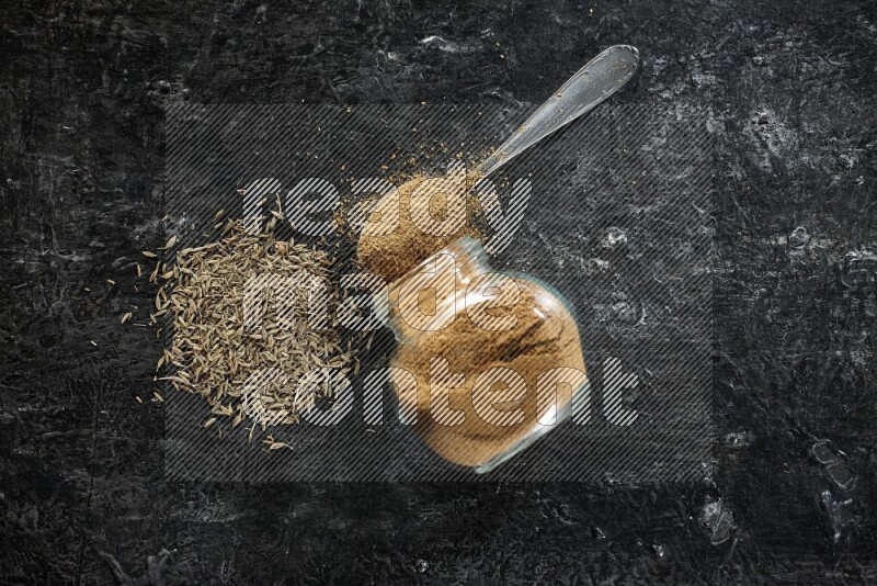 A flipped glass spice jar and a metal spoon full of cumin powder and powder spilled out with cumin seeds on a textured black flooring