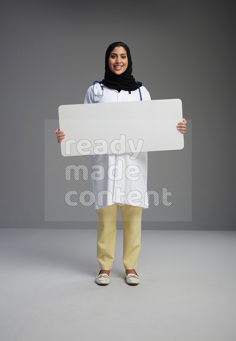 Saudi woman wearing lab coat with stethoscope standing holding board on Gray background