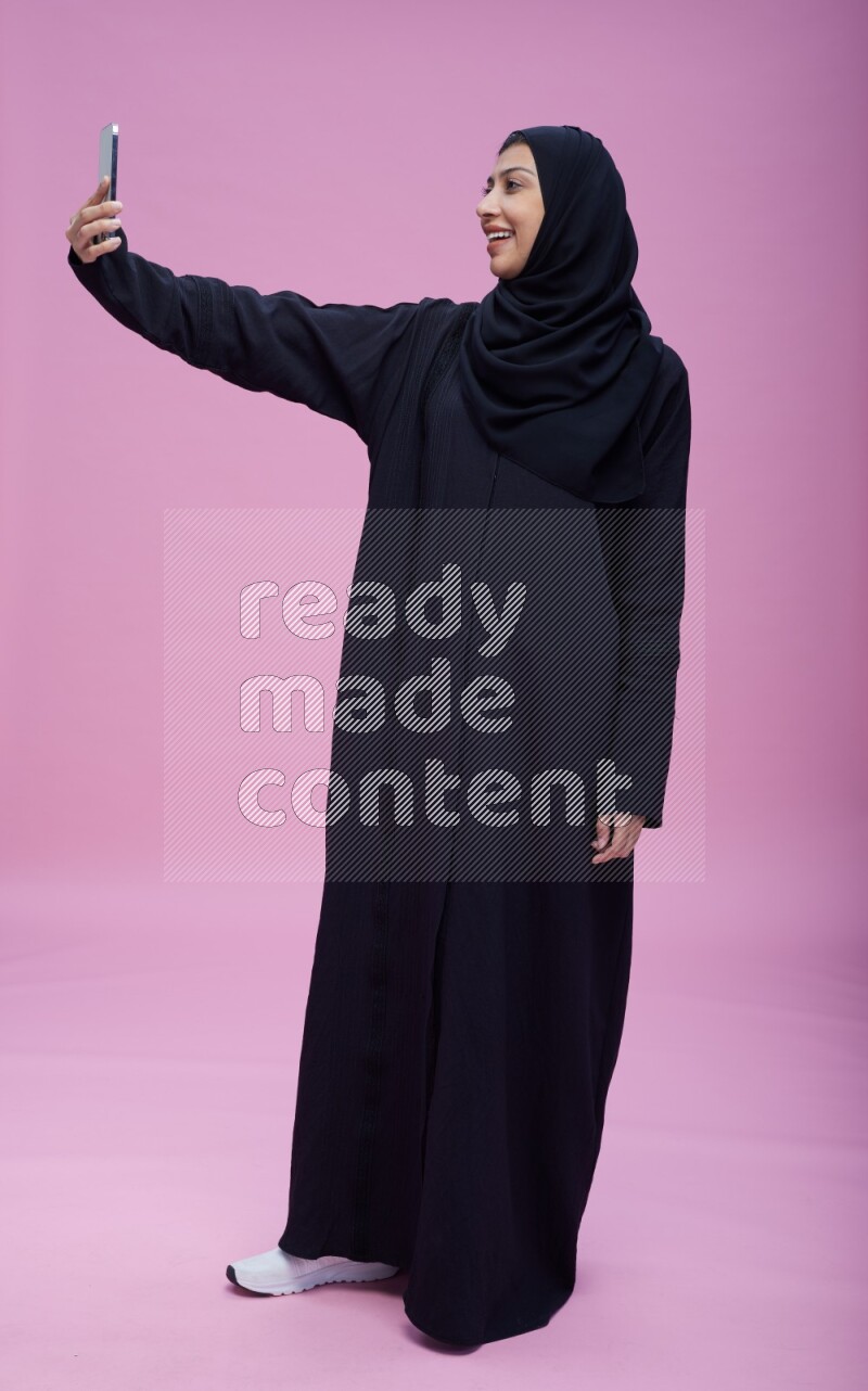 Saudi woman wearing Abaya standing taking selfie on pink background