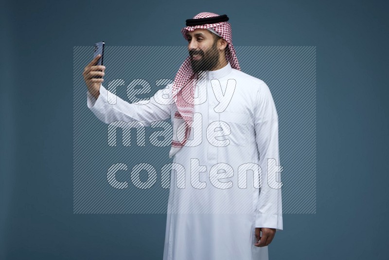 A man Taking Selfie in a blue background wearing Saudi Thob with Shomag