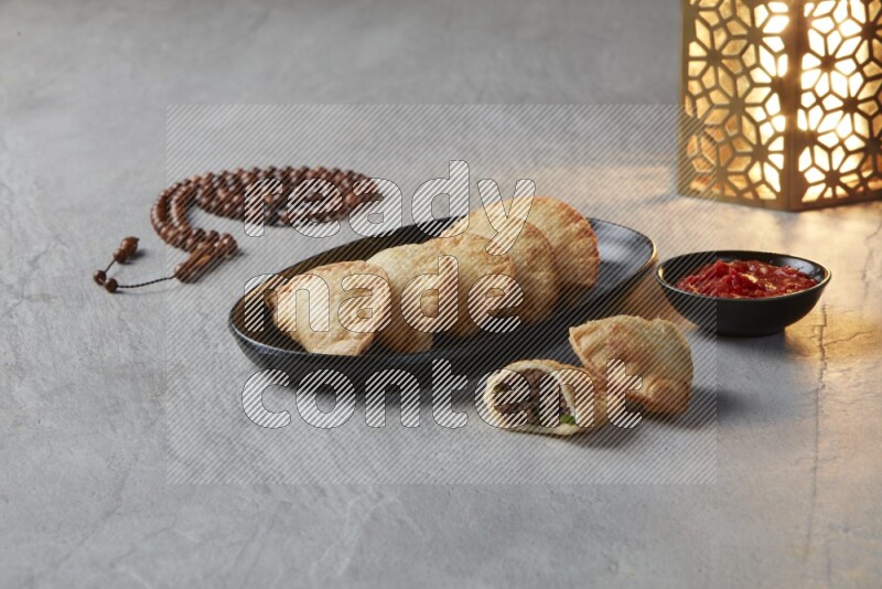 Five fried sambosas in an oval shaped black plate, beside a cut meat sambosa, a brown misbaha and a golden lantern on a gray background