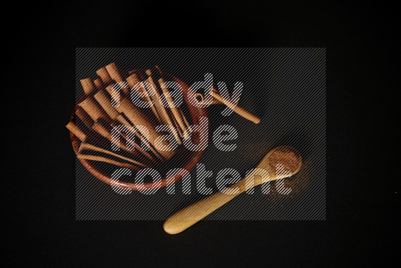 Cinnamon sticks in wooden bowl and cinnamon powder in a wooden spoon on black background