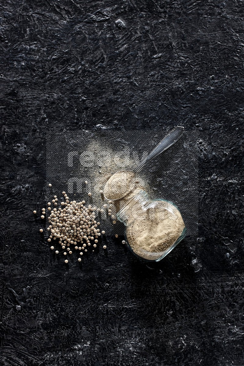 A flipped herbal glass jar and metal spoon full of white pepper powder with spilled powder and pepper beads on textured black flooring