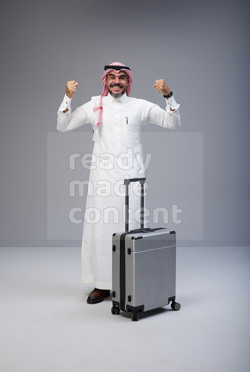 Saudi man wearing Thob and red Shomag standing holding Travel bag on Gray background