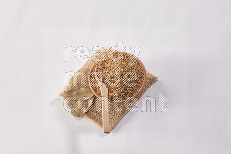 A wooden bowl full of mustard seeds with a wooden spoon on the seed set on burlap fabric on a white flooring
