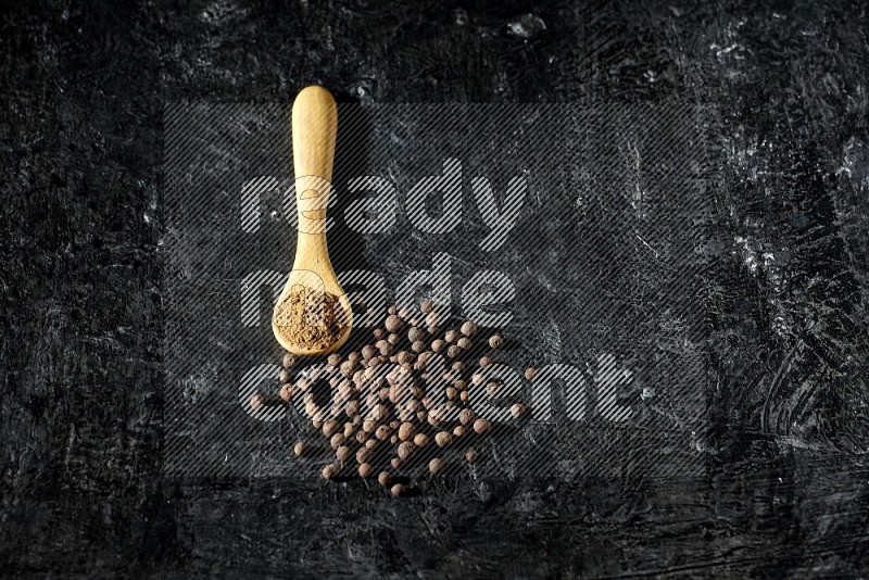 A wooden spoon full of allspice powder and whole balls spreaded on a textured black flooring