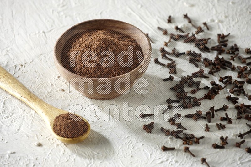 A wooden bowl and wooden spoon full of cloves powder with cloves spread on textured white flooring