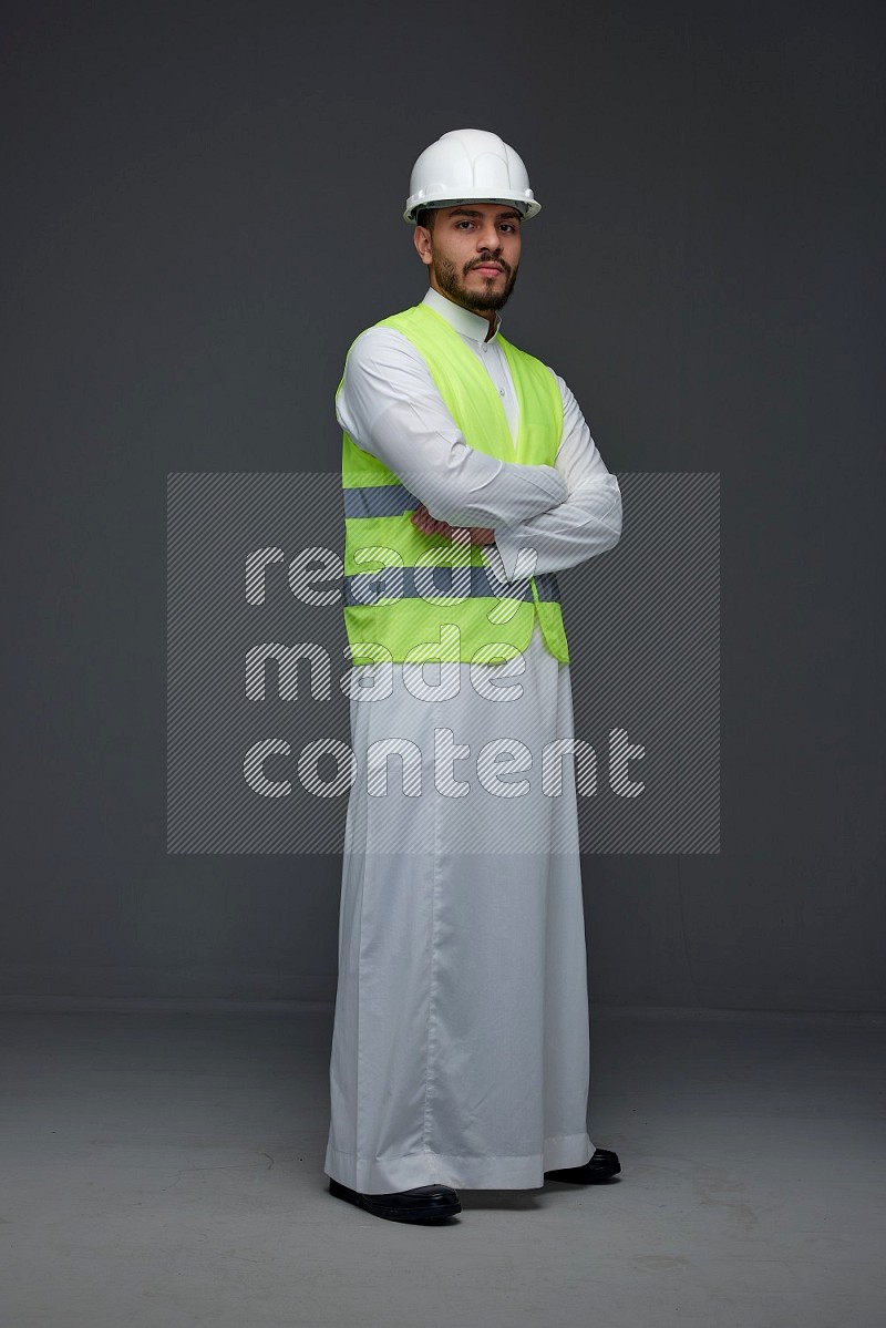 A Saudi man wearing Thobe with a yellow safety vest and white helmet standing and crossing his hands eye level on a gray background