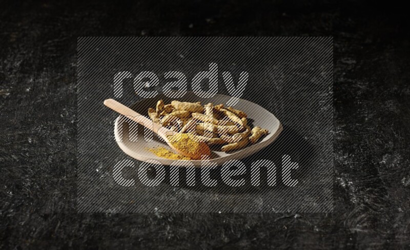 طبق ممتلئ بأصابع الكركم المجففة وملعقة خشبية ممتلئة ببودرة الكركم على خلفية سوداء