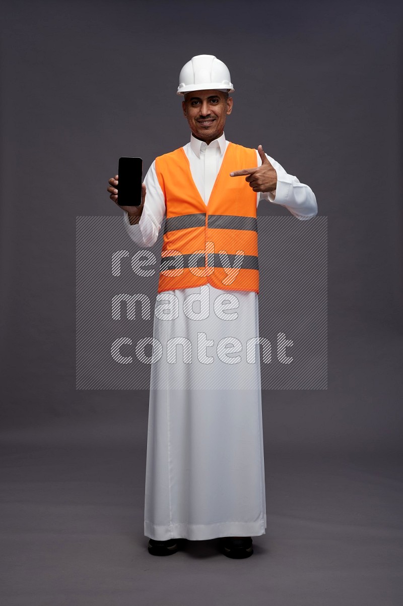 Saudi man wearing thob with engineer vest standing showing phone to camera on gray background