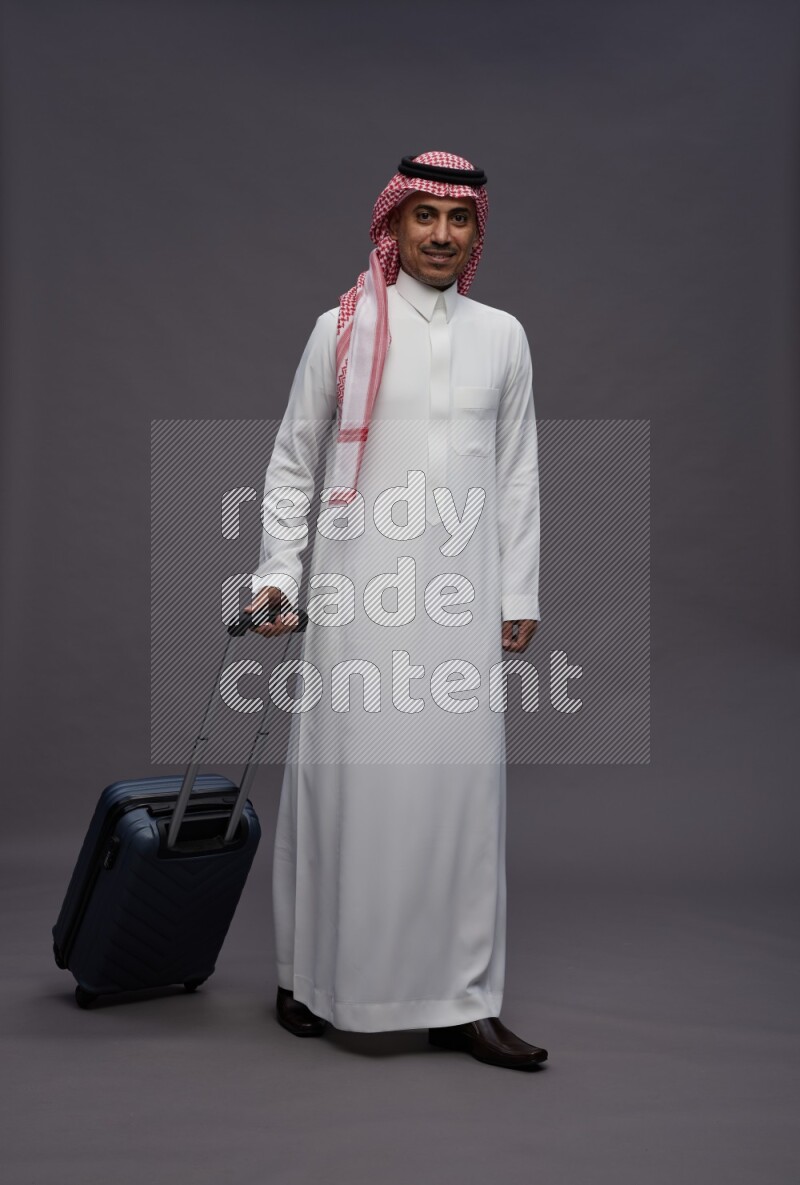 Saudi man wearing thob and shomag standing holding bag on gray background