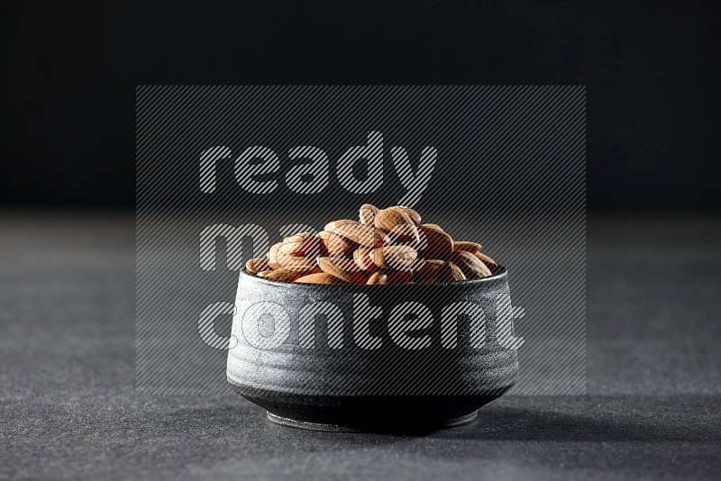 A black pottery bowl full of peeled almonds on a black background in different angles