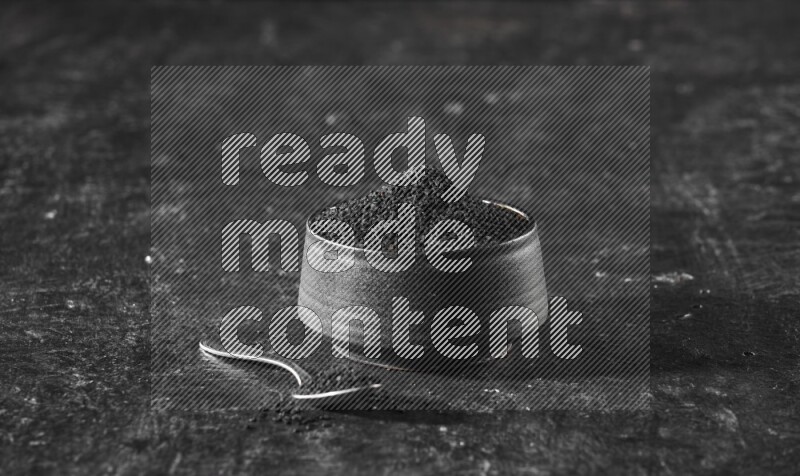A black pottery bowl full of black seeds with a metal spoon full of the seeds on a textured black flooring